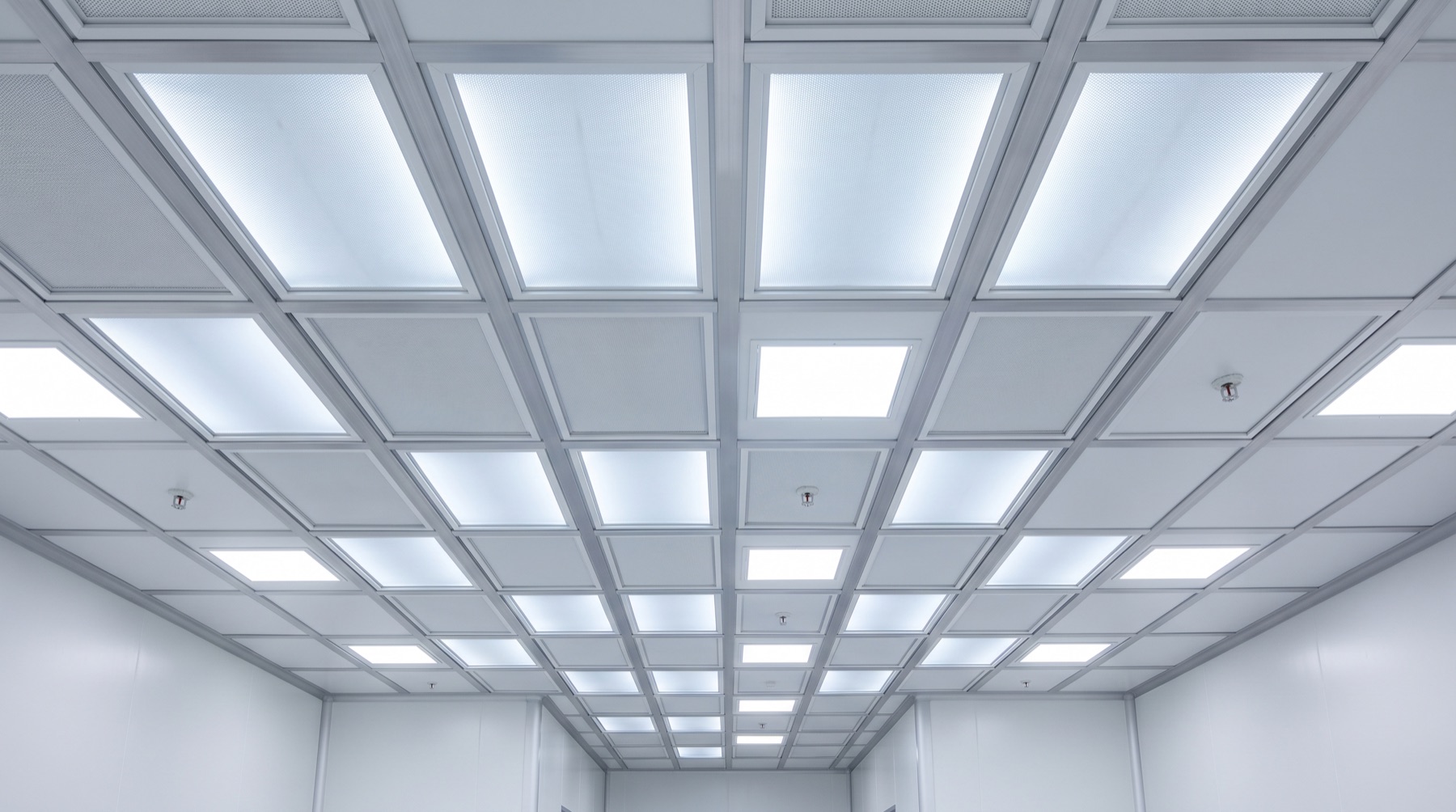 Looking up at a cleanroom ceiling grid filled edge to edge with filter modules, lit from within, above a corridor with a smooth coved floor